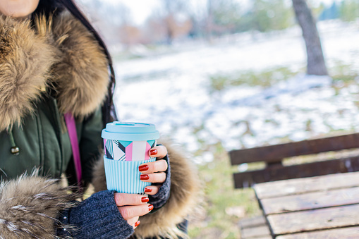 Beautiful young woman is enjoying her cup of coffee in public park on a snowy winter day, Coffee break.