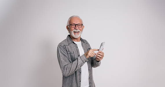 Portrait of happy old male manager using mobile phone and standing confidently on white background