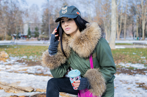Beautiful young woman is enjoying her cup of coffee in public park on a snowy winter day,  Coffee break. She is talking on the phone.