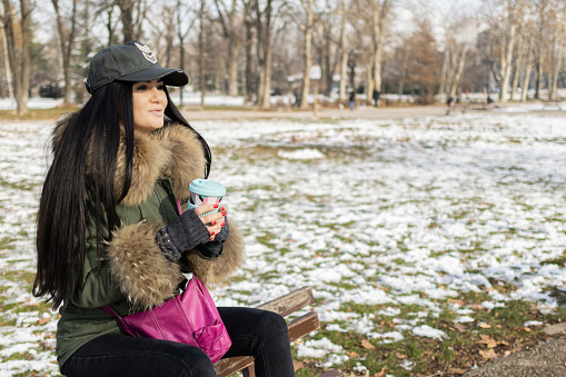 Beautiful young woman is enjoying her cup of coffee in public park on a snowy winter day,  Coffee break.