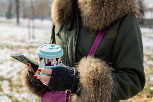 Beautiful young woman is enjoying her cup of coffee in public park on a snowy winter day,  Coffee break. She is taking selfie, or video call.