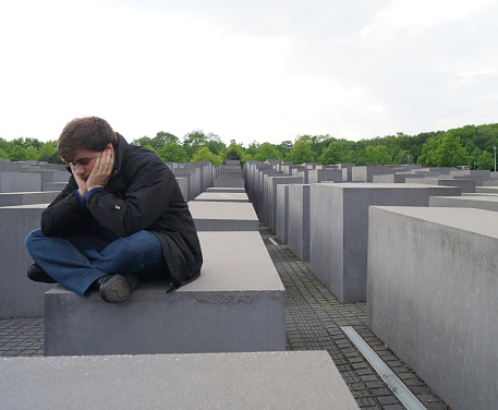 Berlin, Germany; May, 13 - 2017: A young man sits in contemplation on a monolith at the Holocaust Memorial, holding his head with a lost gaze, surrounded by the haunting atmosphere of remembrance