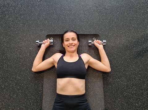 Smiling young woman lifting dumbbells lying on mat. Sports fitness health