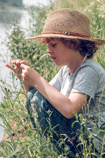 Side view of content kid in straw hat sitting on grass on channel coast and enjoying summer day while playing with grass