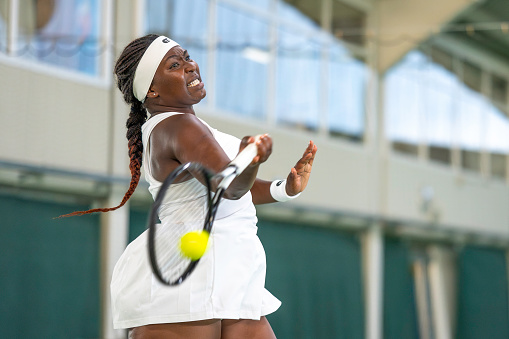 African american female tennis player with tensed facial expression hitting ball while training in court, medium shot
