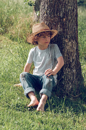 Full length of barefoot pensive kid in straw hat and jeans sitting on green grass leaning on tree and looking away while enjoying summer day