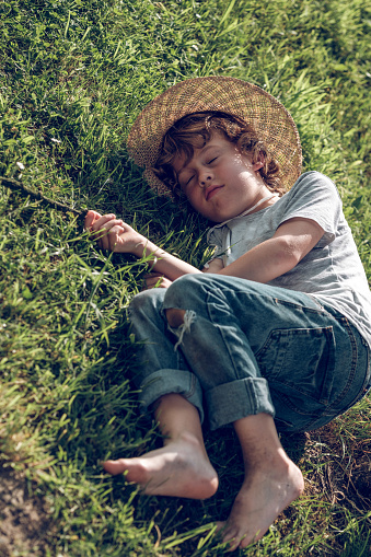From above of full body cute barefoot preteen boy in wicker hat and jeans lying on green grass and falling asleep in sunlight