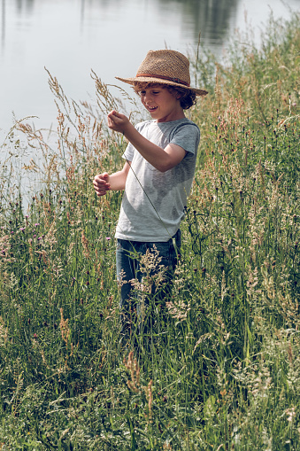 Cheerful kid in straw hat and jeans playing with tall grass while standing on shore of calm channel in summer day and looking away