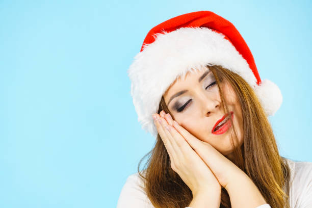 Tired woman wearing santa hat gesturing sleep gesutre with her hands. Adult female being sleepy, ready to take a nap. Christmas time.