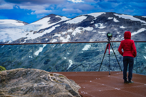Female tourist with camera on tripod taking travel picture, enjoying mountains landscape from Dalsnibba area Geiranger Skywalk viewing platform, Norway.