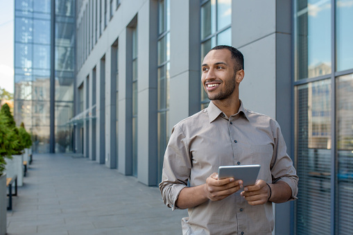 A businessman with a tablet computer in his hands from the outside of an office building, the man is smiling and looking away. An employee in a shirt walks outside an office building, uses an app.