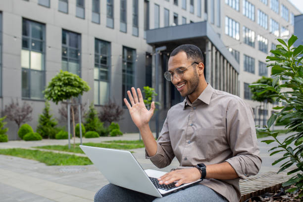 istock Businessman using laptop for video call, man sitting on bench outside office building, waving to interlocutor, greeting gesture 1963091438