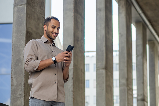 Smiling African American businessman in casual shirt using phone outdoors, possibly texting or browsing.
