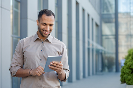 Smiling young man using tablet in urban setting, portraying connectivity, technology, and casual business outdoors.