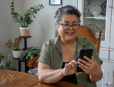 Latina woman wearing glasses and scrolling on smartphone in dining room with plants in background, with copy space