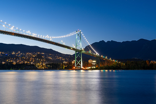 A long exposure photo during blue hour of Lions Gate Bridge in Vancouver, Brithish Columbia with North Vancouver in the background as viewed from Stanley Park.