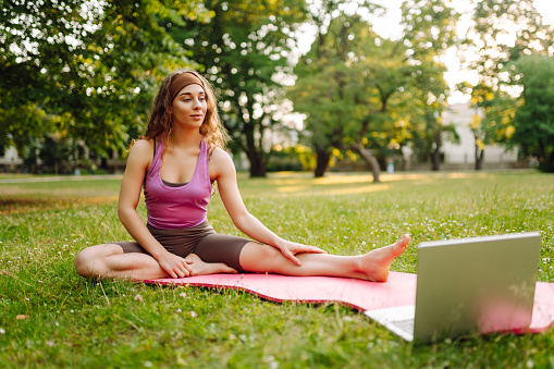 Young woman doing fitness stretching yoga exercise in the park while using laptop for online class or virtual tutorials. Training workout. Healthy lifestyle, sport. Online training, course.