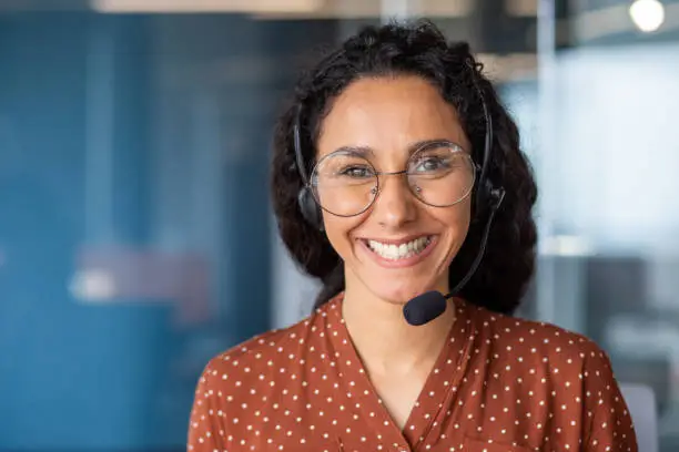 Photo of Close-up portrait of a woman with a headset, an online customer support worker smiling and looking at the camera, a business woman advising clients, working inside the office, using video call