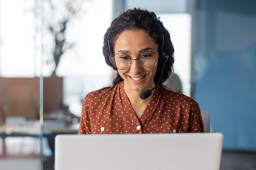 Young beautiful latin american woman with headset phone talking on video call using laptop, business woman inside office building close up, happy customer support tech worker.