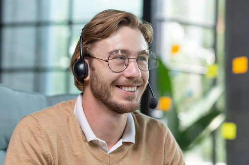 Successful businessman with video call headset working inside office, man consulting online clients, talking with colleagues on online remote meeting, sitting at workplace with laptop