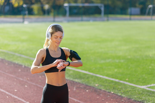 A focused female runner with headphones tunes her workout, checking her smartwatch on a sunny track field.