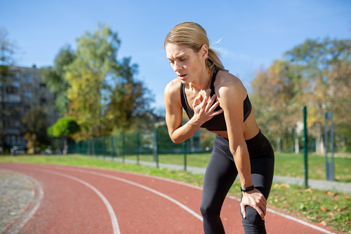 Athletic woman taking a break from a run on a sunny day, holding her knee and catching her breath on a track.