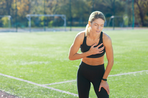 Determined woman in sportswear taking a break, catching her breath post-workout on a sunny sports field.