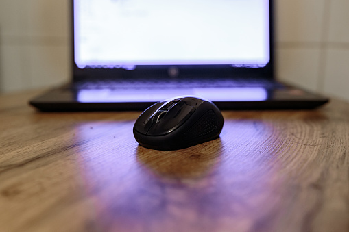 A black wireless computer mouse in front of a laptop pc with bright display