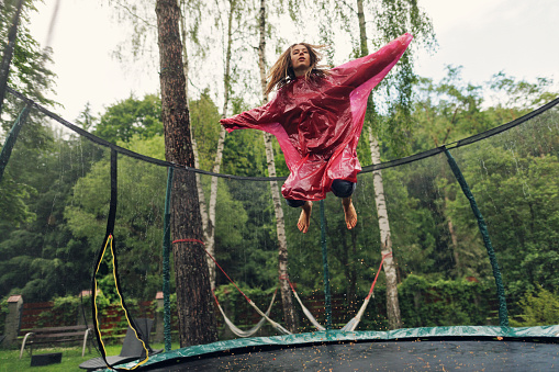 Teenage girl wearing raincoat is jumping on trampoline on a rainy day\nSlow motion footage from Canon R5