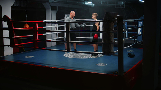 Boy in boxing gloves stands on ring and talks with trainer. Adult man consults young boxer and explains fighting techniques. Teen prepares to fighting match in dark gym. Physical activity and workout.