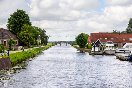 Canal through a small town in the countryside of Netherland on an overcast summer day. Motorboats moored to jetties are in foreground while a wind farm is visible in distance.