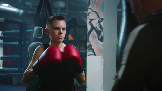 Confident young fighter hits punching bags while training in dark boxing gym. Athletic boy in boxing gloves exercises with trainer before fighting tournament. Physical activity and intensive workout.