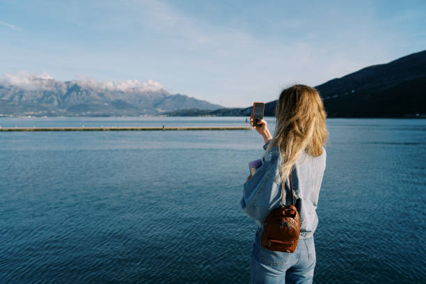 istock Woman shoots a sea and a mountain range with a smartphone while standing on the shore. Back view 1963560037