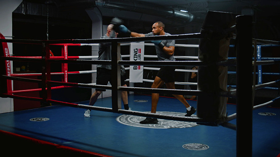 African American fighter exercises and prepares for fighting match with Caucasian trainer on boxing ring. Athletic man in boxing gloves hits punching mitts in dark gym. Physical activity and workout.