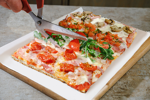 Close-up on pizza with various juicy toppings on one flatbread next to each other in rectangular stripes, cutting with special scissors and serving pizza al taglio in fast food