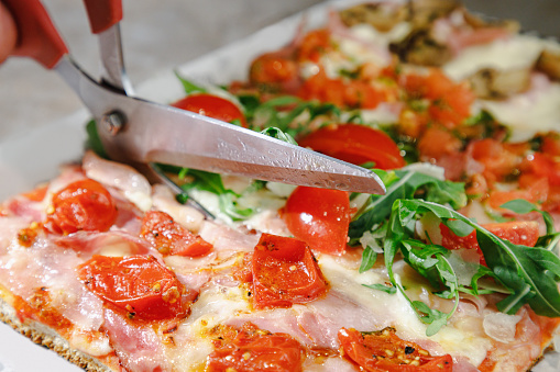 Close-up on hand cutting pizza with various juicy toppings on one flatbread next to each other in rectangular stripes using special scissors, fast food pizza al taglio