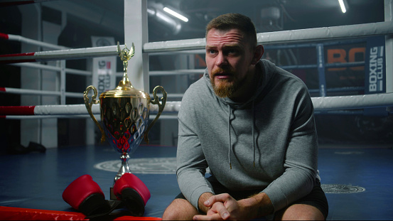 Male boxer sits near boxing ring and discusses victory in tournament with interviewer. Athlete talks after championship or competition in boxing gym. Cup of winner and boxing gloves lies near fighter.