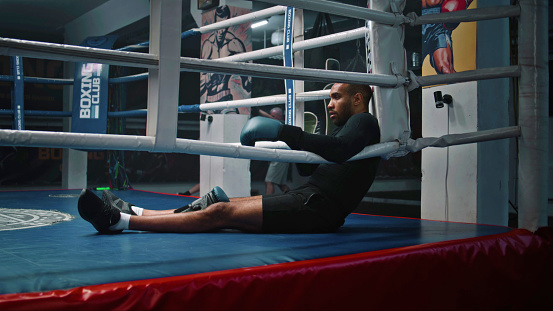 Tired and exhausted African American fighter sits in boxing ring corner. Male boxer takes off his boxing gloves and takes break during training. Athlete prepares to fight or competition in boxing gym.