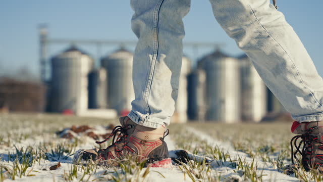 Low Section Side View of Female Farmer in Boots Walking on Snowy Field with Silos in Background on Sunny Day