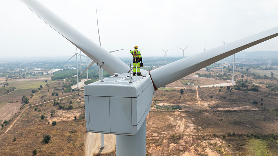 Specialist windmill engineer with green safety jacket and full PPE include safety harness work on top of wind turbine