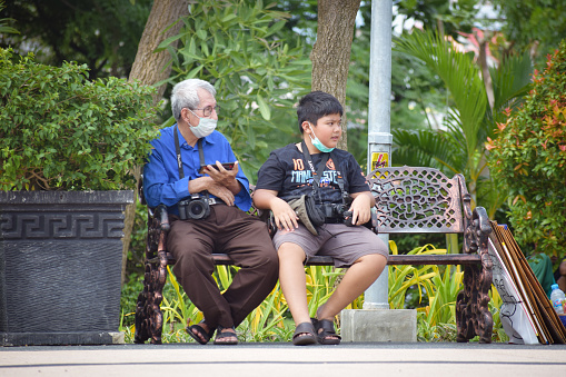 10/05/2023 surabaya indonesia at public park asian old & young photographer sit on bench see at one direction with masked on