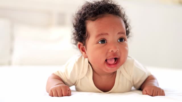 laughing portrait close-up of a small African-American baby girl in a white bodysuit on the bed at home, a funny six-month-old smiling and joyful black newborn baby lying on her tummy
