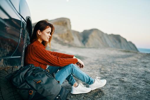 woman in jeans and sneakers sits on the sand near the car in nature. High quality photo