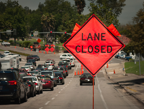 An orange diamond shaped roadwork sign stating lane closed with traffic backed up in the distance