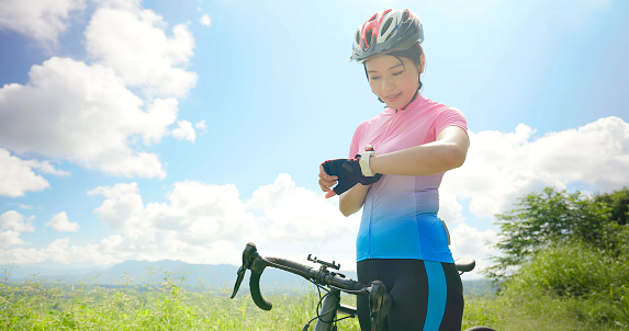 enjoy sport or healthy lifestyle concept - asian young woman standing by a bicycle on the road wearing helmet is checking her smart watch and ready to ride