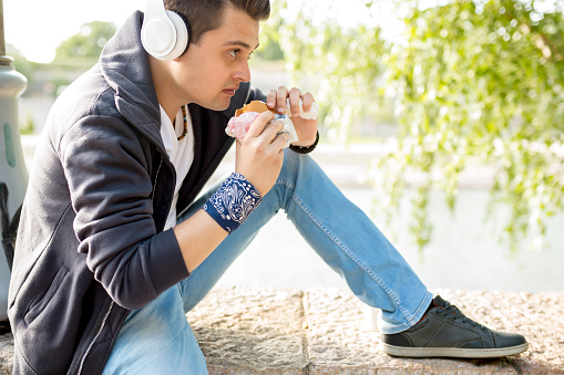 Stylish guy sits on the quay on the background of the river and the bridge. He wears blue jeans. Male looks up. Outdoors. Horizontal.