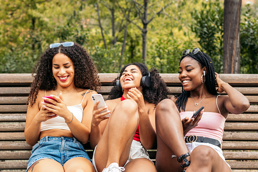 African american female friends sitting on a park bench laughing and having fun while using smartphone and listening to music with headphones.