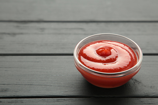 Small glass condiment bowl of red tomato sauce ketchup on black background. Top view