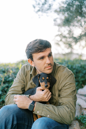 Pensive guy sits in a clearing with a puppy on his knees, put his chin on his head. High quality photo