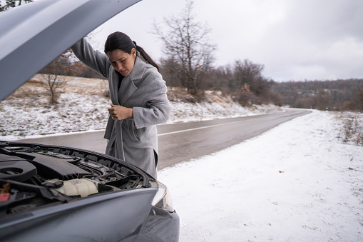 Woman in grey coat opening the car's bonnet and looking under the hood in countryside on cold winter day